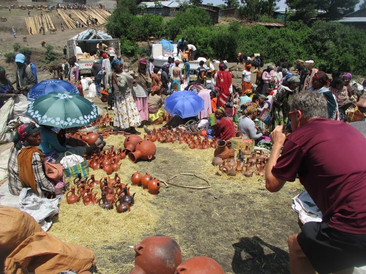 A tourist photographing traditional Ethiopian clay pots and coffee ceremony sets at a lively local market near the Adadi Mariam rock church